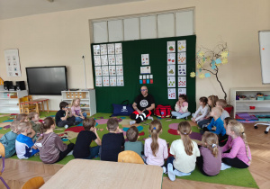 Dzieci siedzą na dywanie i słuchają prowadzącego